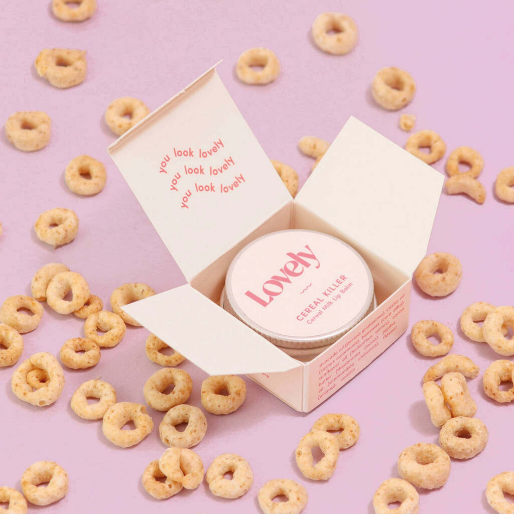 Cereal Milk Cereal Killer Lip Balm 15ml in open box surrounded by cereal loops on pink background.