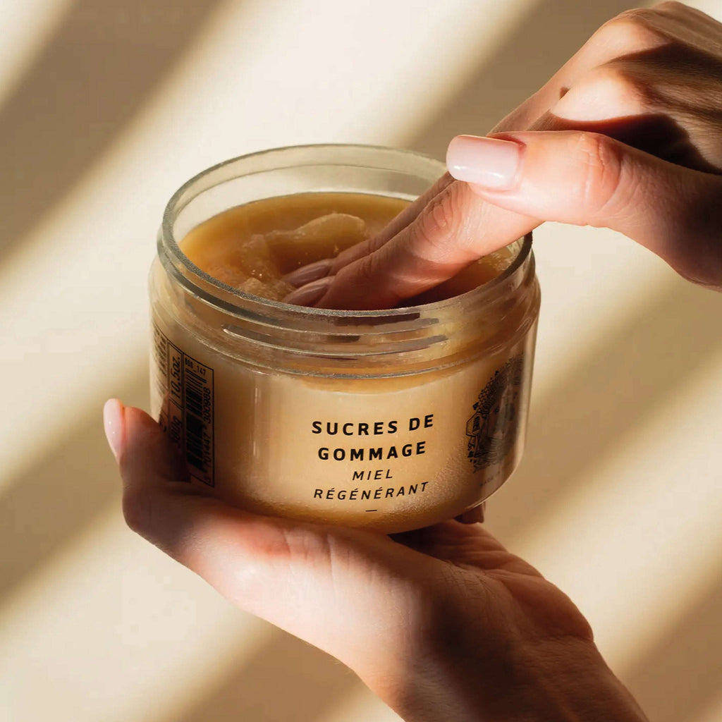 Panier Des Sens Honey Body Sugar Scrub 300g in a jar being scooped by hand, featuring natural exfoliating sugar flakes.