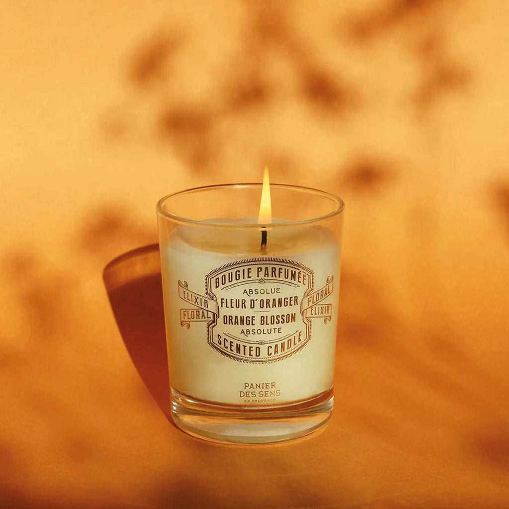 Panier Des Sens Orange Blossom Scented Candle 180g with glowing flame, exuding citrus freshness in a sunny setting.